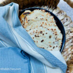 Four flat bread on a black plate, covered in a white and blue cloth and a solid blue cloth, placed in a wicker basket.