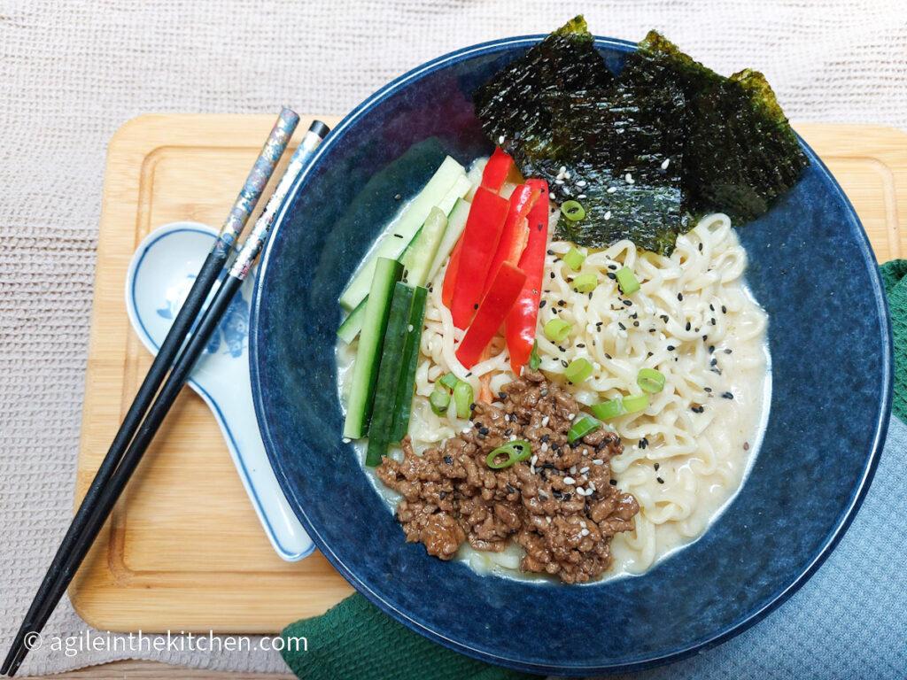 Creamy Ramen in a blue bowl, topped with Asian mince, sliced cucumbers and red peppers, nori sheets, sprinkled with green onions and black sesame seeds. Next to the bowl is a pair of black chopsticks, a large ceramic spoon all on top of a wooden chopping board.