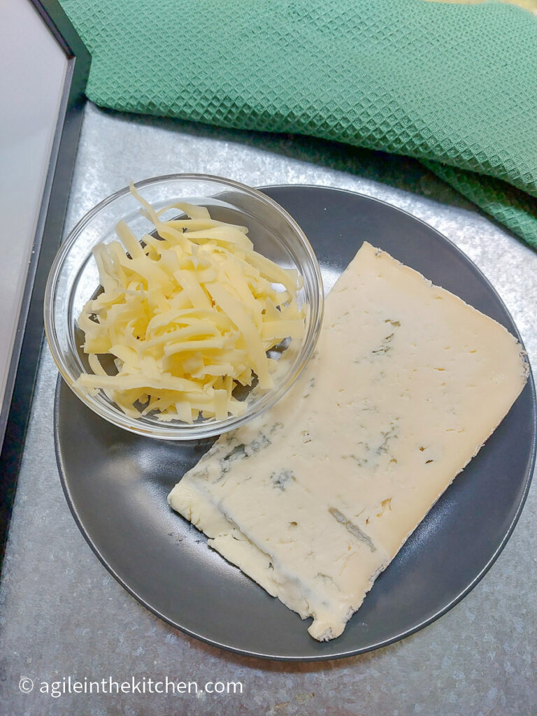 On a grey plate, a glass bowl of shredded cheddar cheese and a wedge of blue cheese