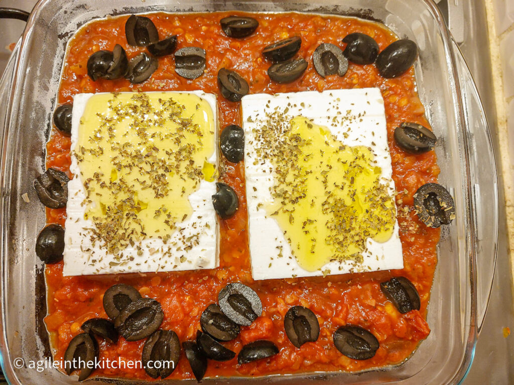 A oven safe glass, square pan with tomato sauce, halved black olives, two bricks of feta cheese, drizzled olive oil and sprinkled oregano