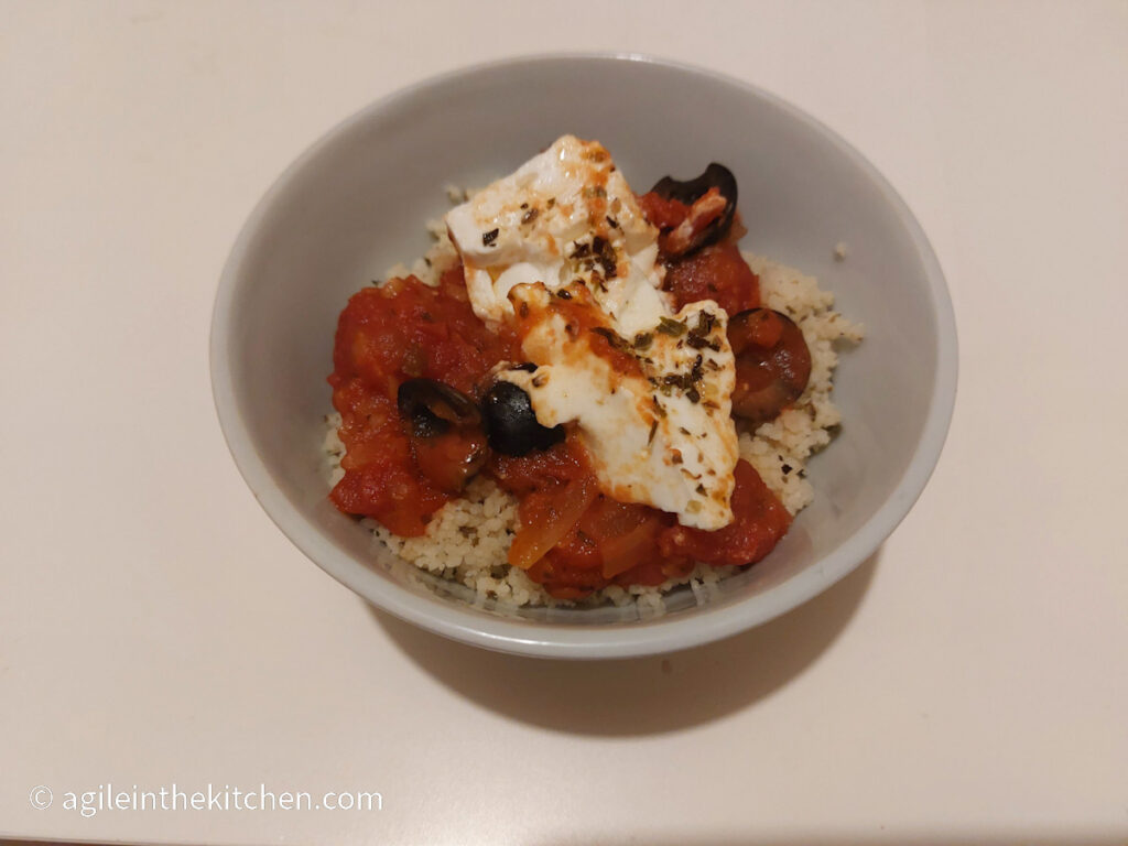 A white bowl with couscous and feta cheese bake, tomato sauce and black olives