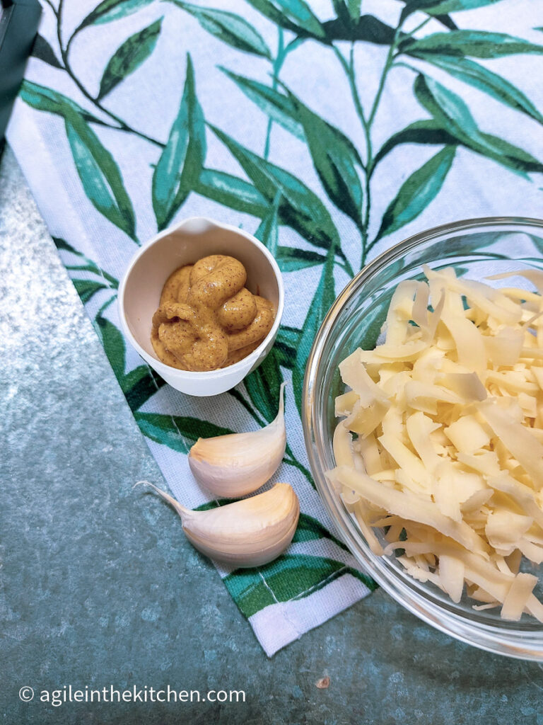 On a silver metal background, a white tea towel with green leaves. On top of the tea towel two cloves of garlic, a small white container with Dijon mustard and a clear glass bowl with shredded cheese.