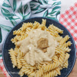 On a wooden background, a red checkered tea towel and a white tea towel with green leaves, sitting on top of them is a blue shallow bowl with fusili pasta topped with Dijon bechamel sauce and a dollop of Dijon mustard.