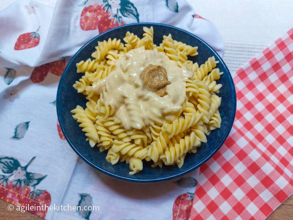 Mac n' Cheese pasta and sauce with a dollop of Dijon mustard on top in a blue bowl. The bowl is resting on a tablecloth with strawberries and a red gingham cloth.