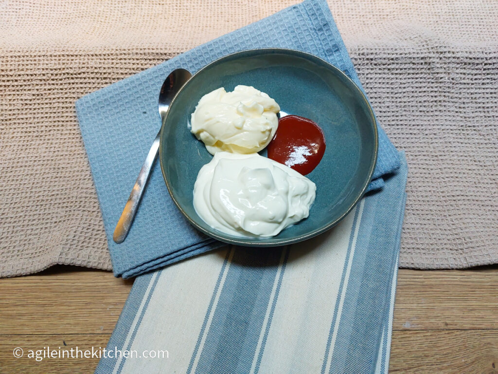 A blue plate with three dollops of Greek yogurt, mayonnaise, and siracha, with a spoon resting on the side. Sitting on top of one blue napkin and one blue striped napkin. In the background a beige textured table cloth.