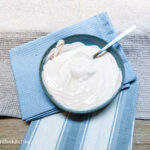 A blue plate with 1-2-3 dressing mixed together with a spoon stuck in. Resting on the side. Sitting on top of one blue napkin and one blue striped napkin. In the background a beige textured table cloth.