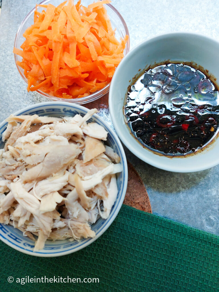 On a silver metal background and a green table cloth there are three bowls of similar size, one with shredded chicken, one with shredded carrot and one with a soy sauce based dressing