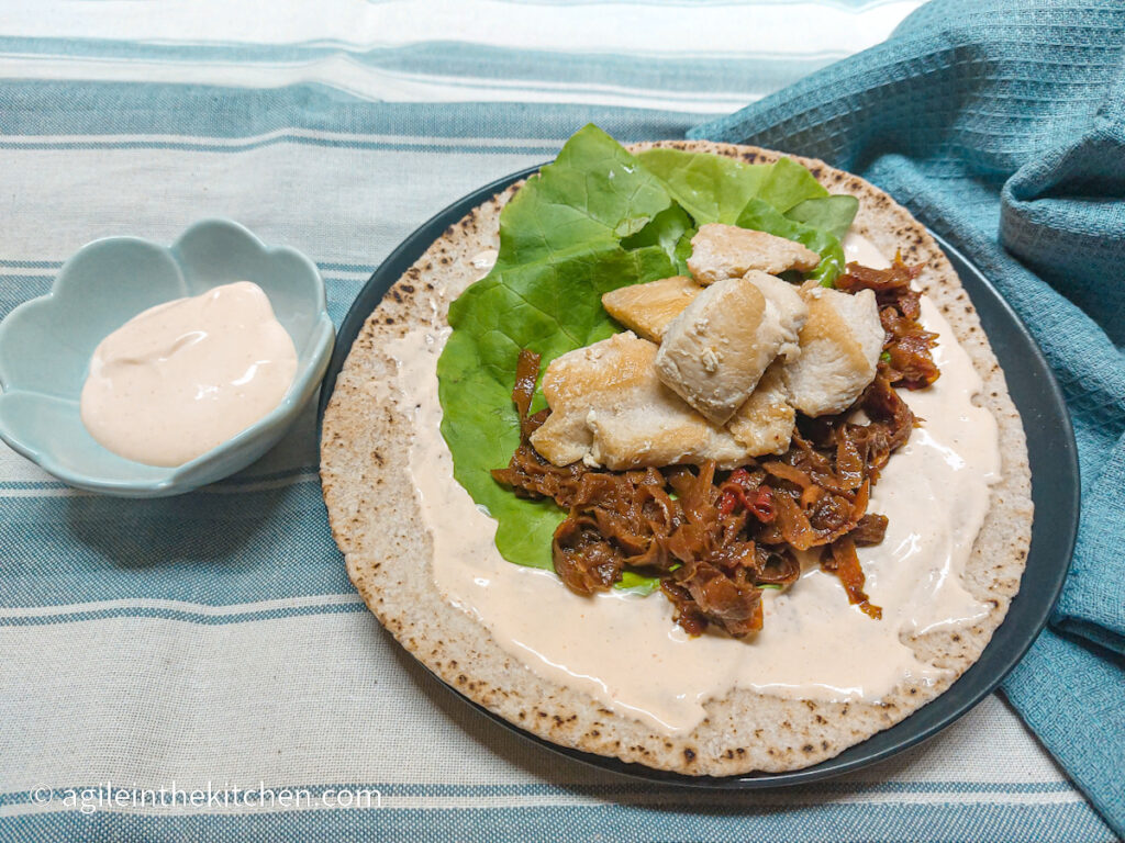 On a blue and white horizontal striped background, from the left, a flower shaped bowl with dressing, a plate with an Asian chicken wrap with green lettuce and diced chicken and a blue napkin crumbled up.