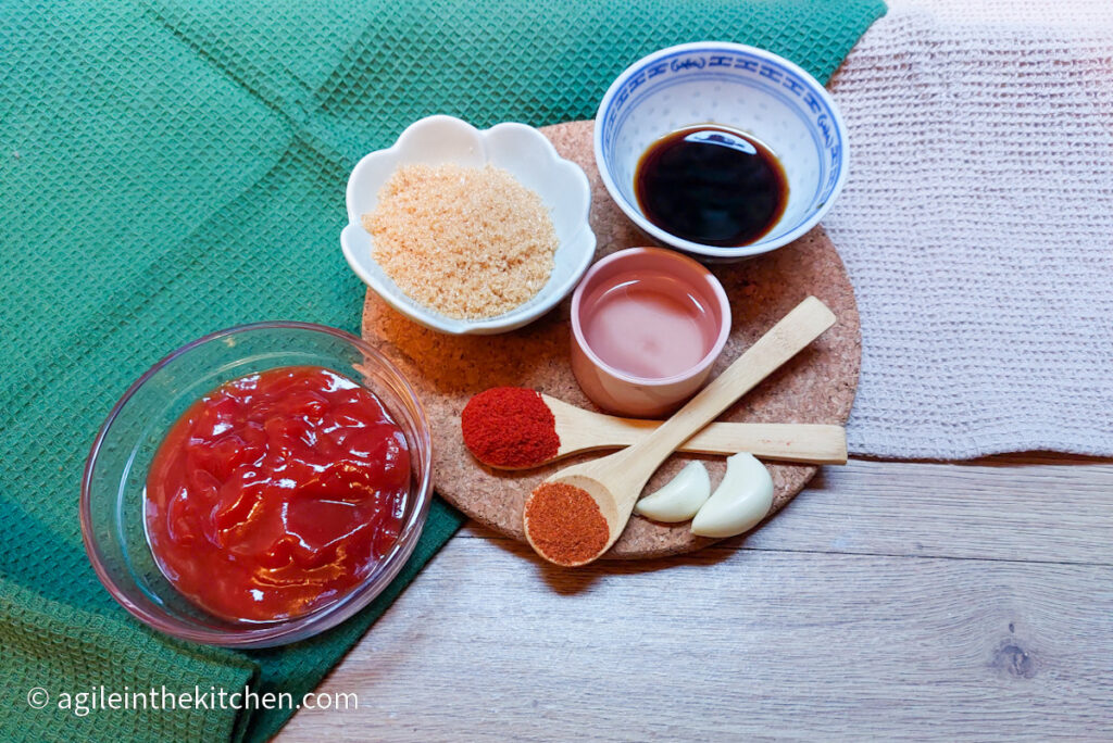 On a green and beige table cloth on top of a wooden table, the ingredients to make barbecue sauce, from the left: ketchup in a glass bowl, brown sugar in a bowl, two wooden teaspoons with paprika and chili powder, a bowl of white vinegar, two garlic cloves and a bowl of soy sauce.