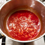 A red barbecue sauce is cooking in a silver coloured pot on a gas stove top