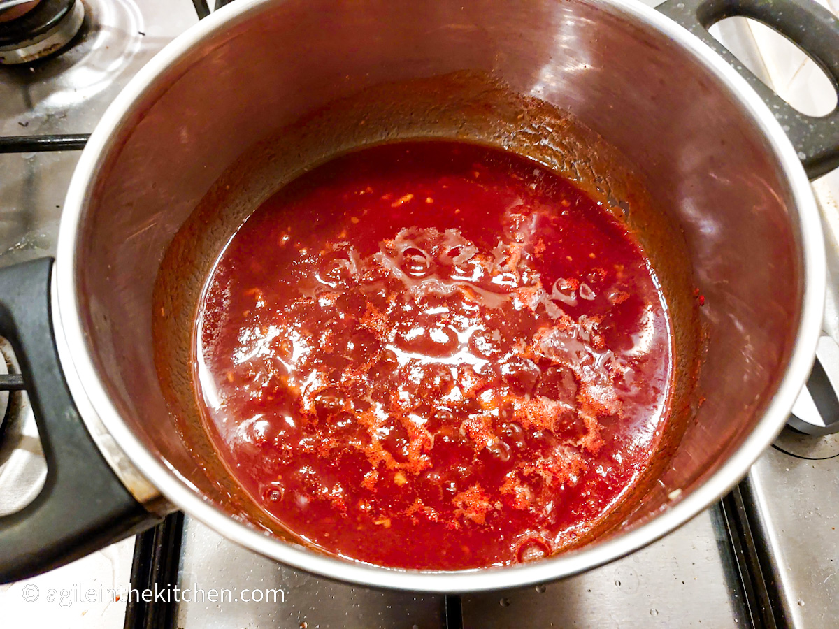 A red barbecue sauce is cooking in a silver coloured pot on a gas stove top