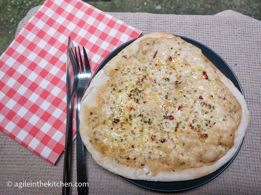 A pizza bianca sitting on a black plate on a beige, textured table cloth and a folded red checkered napkin to the left, with a knife and fork on it.