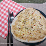 A pizza bianca sitting on a black plate on a beige, textured table cloth and a folded red checkered napkin to the left, with a knife and fork on it.