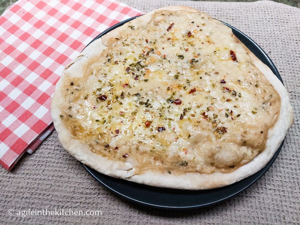 A pizza bianca sitting on a black plate on a beige, textured table cloth and a folded red checkered napkin to the left.