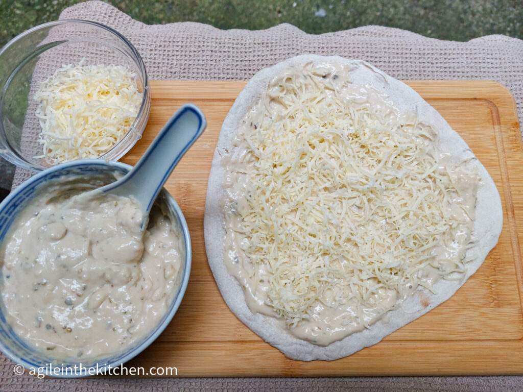 A pizza bianca being prepared with bechamel sauce and shredded cheese, sitting on a wooden cutting board on a beige, textured table cloth. To the left is a bowl of bechamel sauce and a bowl of shredded cheese.