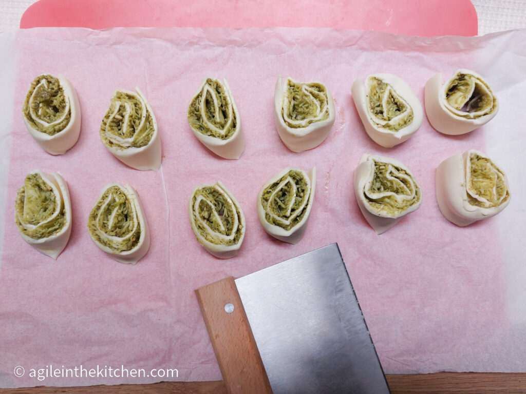 On a white textured table cloth, a red plastic cutting board, a parchment paper, twelve pesto pinwheels, cut side up, and a dough cutter.