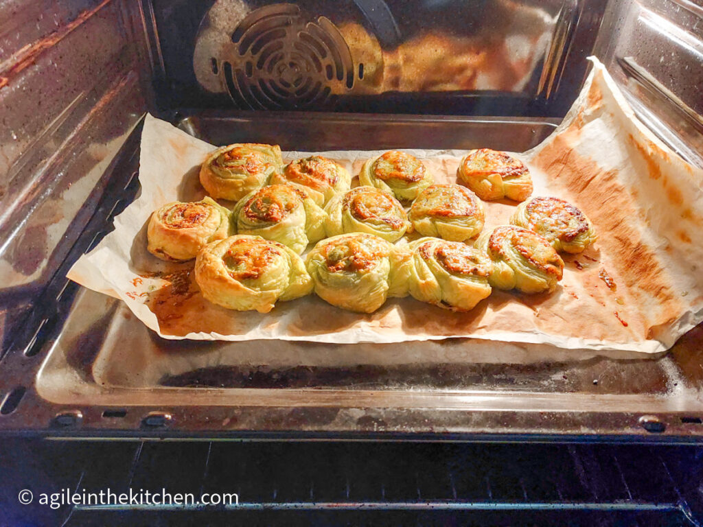 In the oven on a cookie sheet with baking paper, green pesto pinwheels are cooking