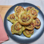 On a white background, a wooden cutting board with a folded peach coloured napkin. A black matte plate with a pile of green pesto pinwheel straight from the oven