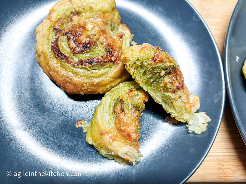 A wooden cutting board with a black, matte plate with two puff pastry pinwheels with green pesto, one is cut in half to show the inside.