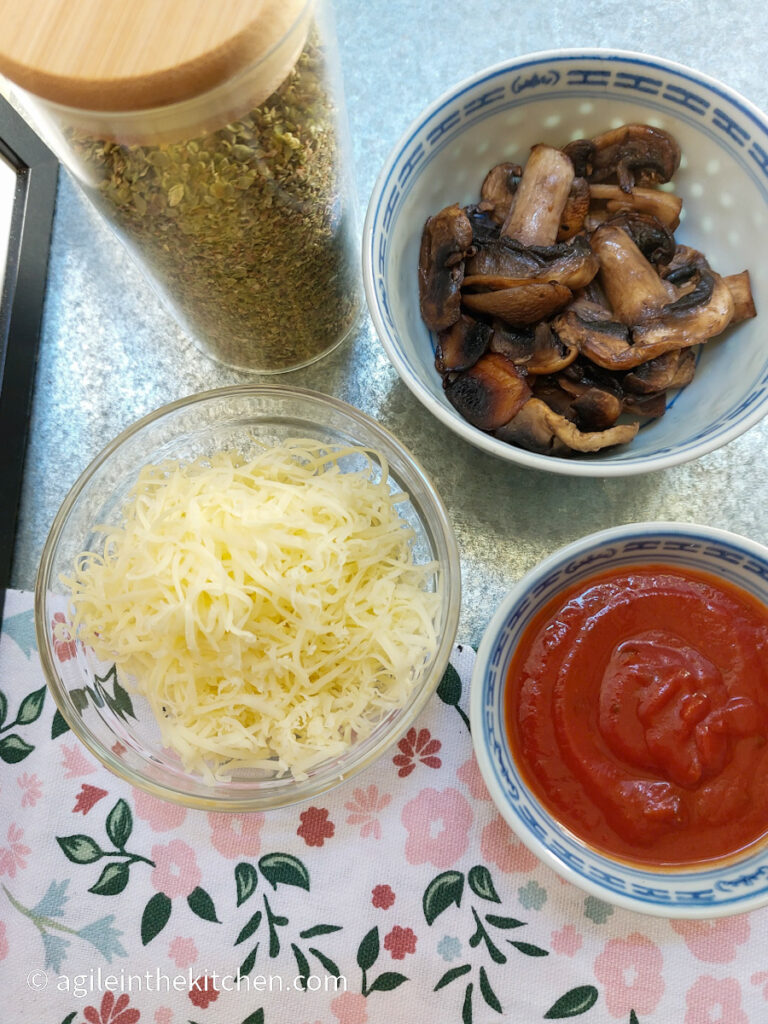 On a silver metal background with a pink flowery table cloth in the bottom half, pizza toppings are laid out. Clockwise, oregano in a glass jar with a wooden lid, fried mushrooms in a bowl, marinara sauce in a smaller bowl, shredded cheese in a glass bowl.