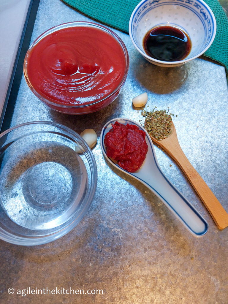 On a silver metal background, ingredients to make a marinara sauce is laid out. Clockwise from the top, tomato passata in a glass bowl, soy sauce in a smaller bowl, two cloves of garlic, one wooden teaspoon with oregano, one spoon with tomato pure and a glass bowl with water.