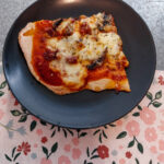 A quarter wedge of pizza with marinara sauce, mushroom and cheese on a black plate. Sitting on a silver metal table with a pink flowery table cloth.