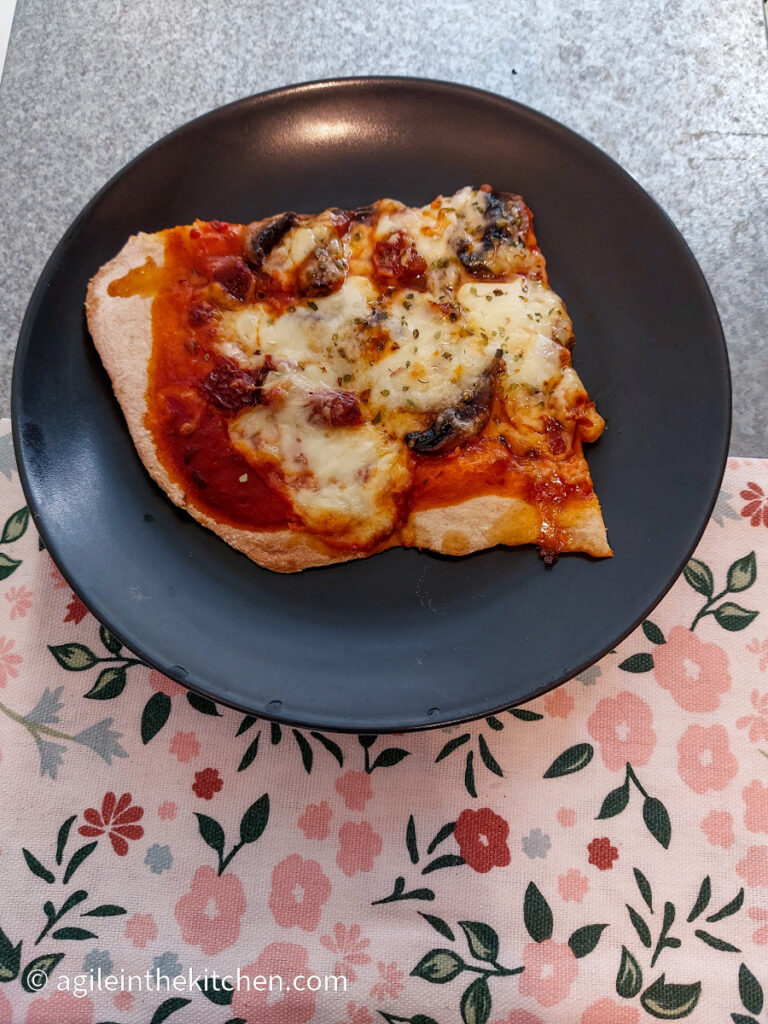 A quarter wedge of pizza with marinara sauce, mushroom and cheese on a black plate. Sitting on a silver metal table with a pink flowery table cloth.
