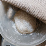 In a white bowl a roundel of pizza dough lays to rest with a beige tea towel covering half the bowl