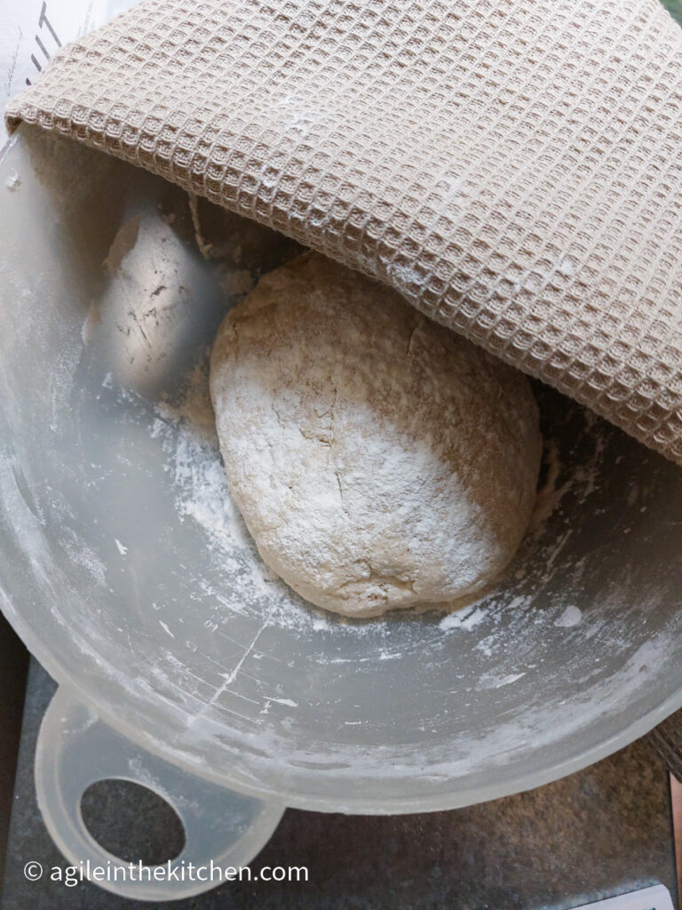 In a white bowl a roundel of pizza dough lays to rest with a beige tea towel covering half the bowl