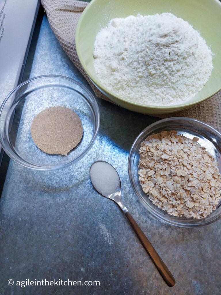 On a silver metal background, clockwise, dry yeast in a glass bowl, all purpose flour in a green bowl, rolled oats in a glass bowl and a teaspoon with salt.