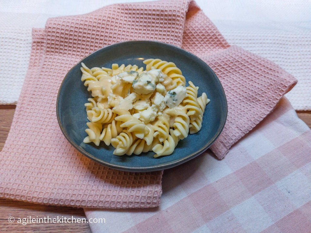 On a background of two pink napkins, one solid pink and one checkered pink, a blue plate with blue cheese mac n' cheese.