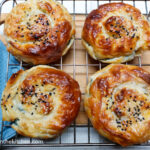 On a wooden cutting board, a wire rack with four Börek taken out of the oven and resting. To the left, a blue napkin.