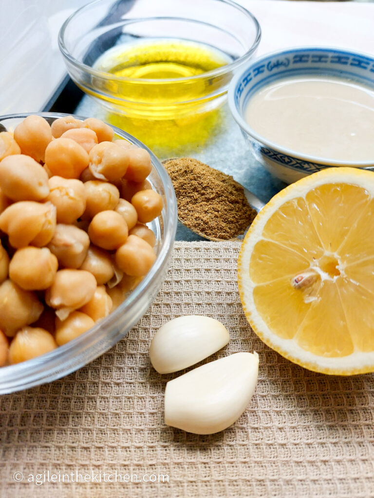 On a background of beige tablecloth, the ingredients to make hummus, clockwise from the top, a glass bowl of olive oil, a bowl of tahini, a lemon cut in half, two cloves of garlic, a glass bowl of cooked chickpeas and a spoonful of cumin.