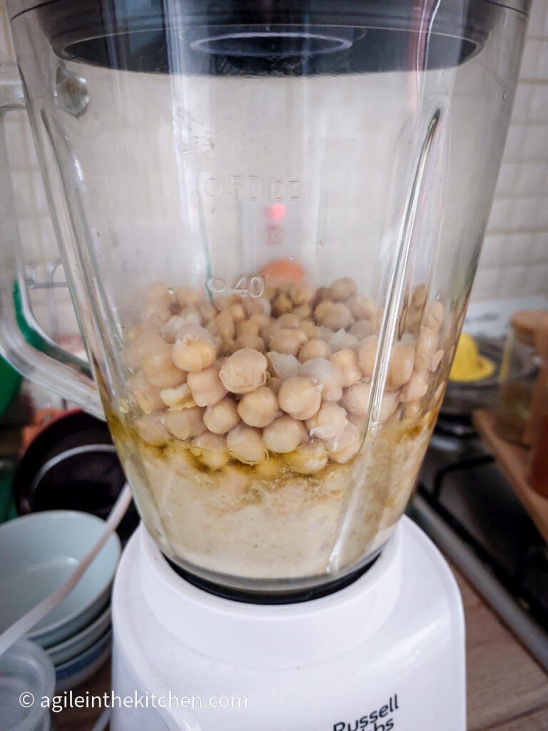 In a standing mixer with a white base and glass top, the ingredients to make hummus, chickpeas, oil, tahini, lemon juice, garlic