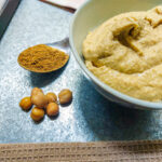 A beige tablecloth covering the lower part of the photo, then on a silver metal background from the left, a spoonful of cumin, a handful of chickpeas and a bowl of hummus