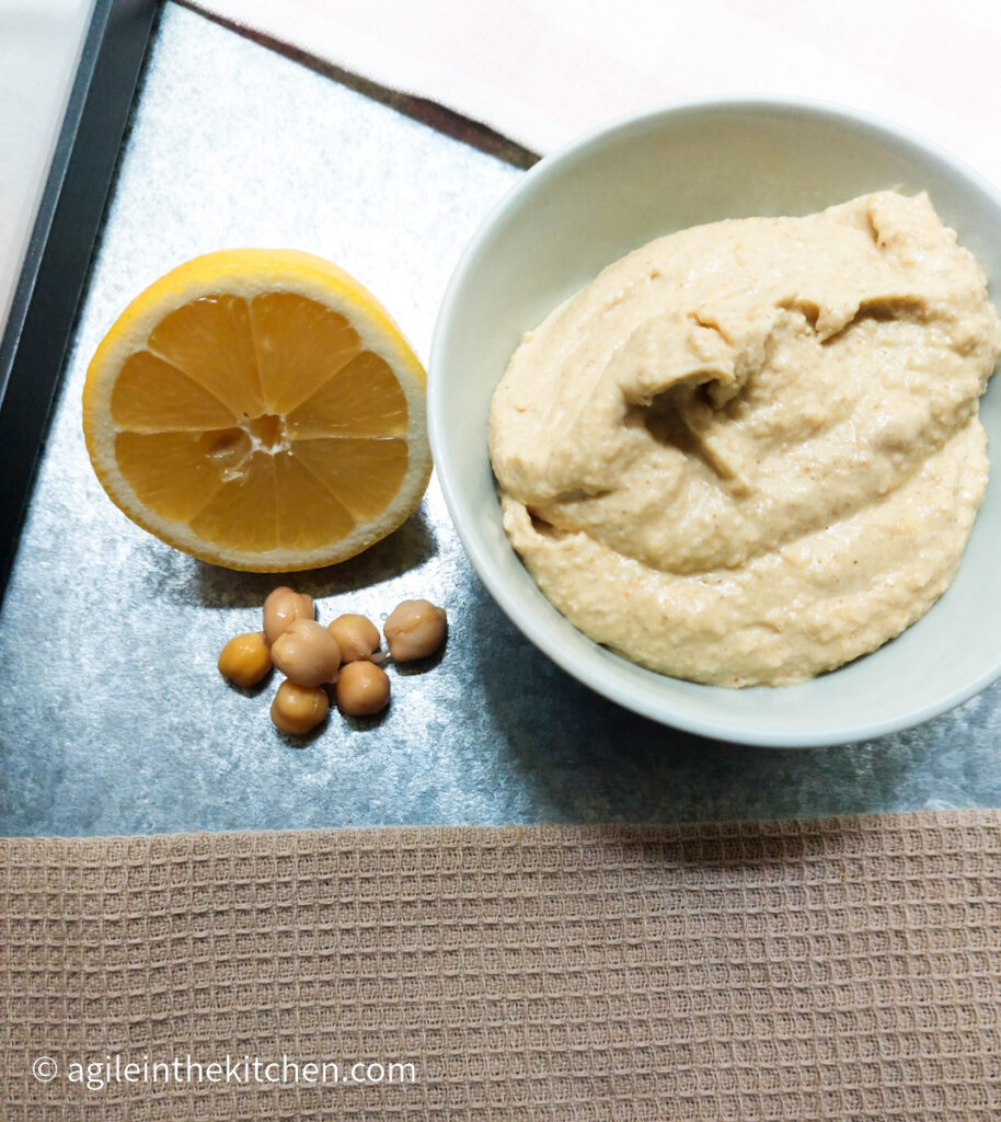 A beige tablecloth covering the lower part of the photo, then on a silver metal background from the left, a lemon cut in half, a handful of chickpeas and a bowl of hummus