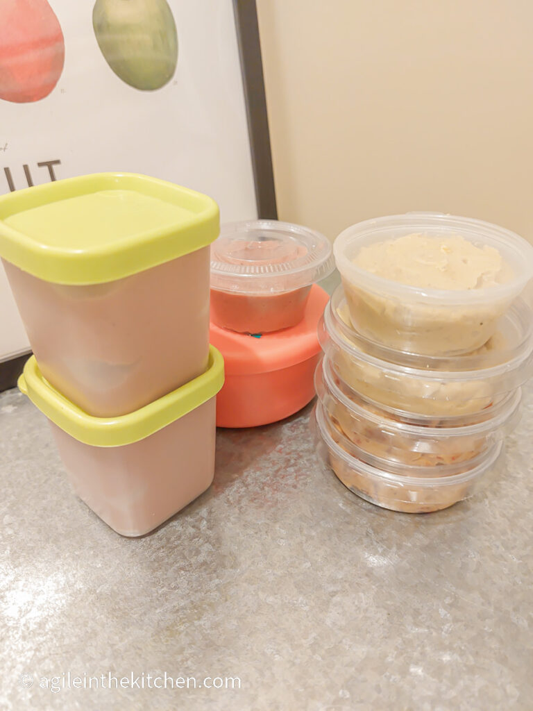 On a silver metal background three stacks of small freezer jars filled with hummus and ready for the freezer.