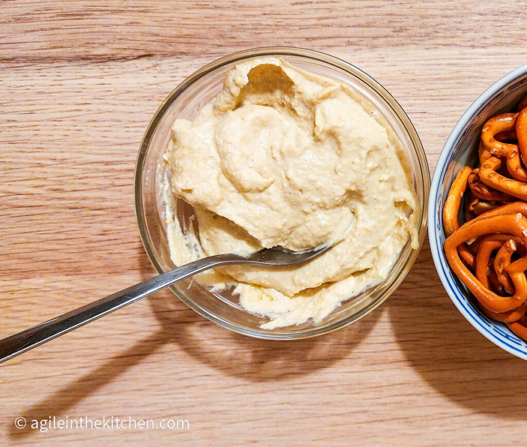 On a wooden background a glass bowl of hummus with a spoon stuck in the bowl, to the left half a bowl of pretzels.