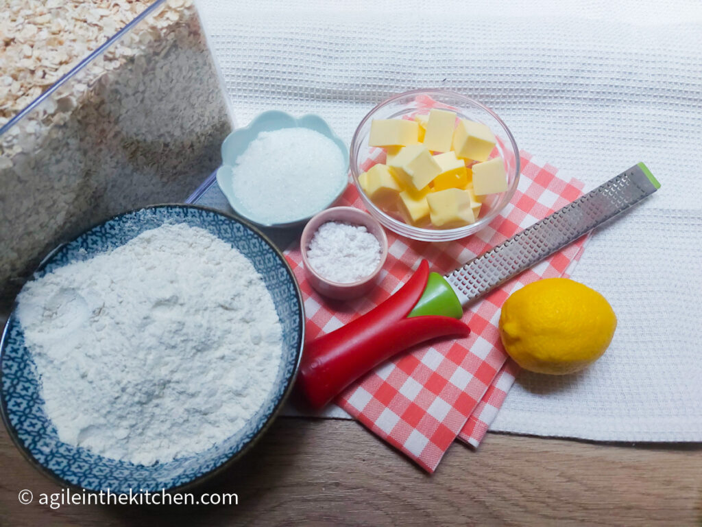 Ingredients to make lemon oat cookies lines up, from the left: a see-through container of oats, a blue bowl of white flour, a light blue flower shaped dish with sugar, a pink small bowl with baking powder, a glass bowl with cut up pieces of butter, a citrus zester with red handle, a lemon. Beneath the ingredients a folded red cloth napkin.