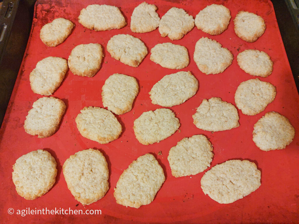 On a red silicone cookie sheet, twenty-four lemon oat cookies have just been taken out of the oven, some of the cookies show slightly brown edges.