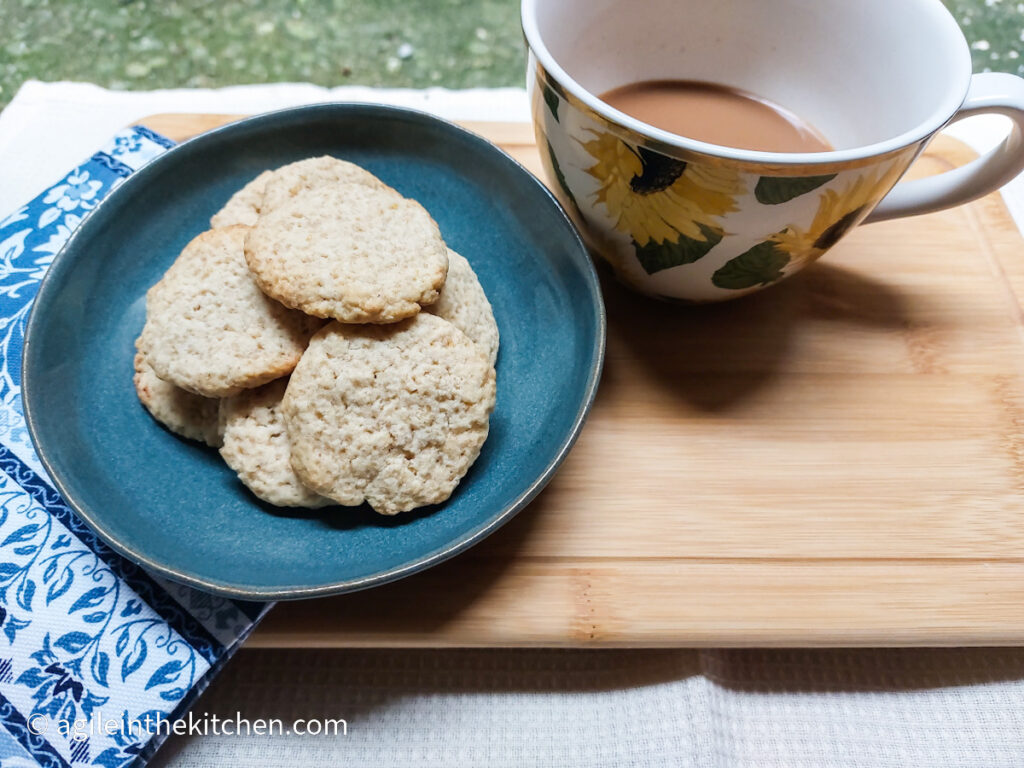 On a wooden cutting board, a blue plate holds a pile of lemon oat cookies, next to the plate is a sunflower printed coffee cup with some milky coffee inside. To the left is a blue patterned napkin.