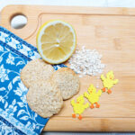 A wooden cutting board, with a folded blue coloured napkin with white flowers in the lower right corner. Three lemon oat cookies are stacked beneath a cut lemon, a handful of oats and to the right, three yellow chicken decorations.