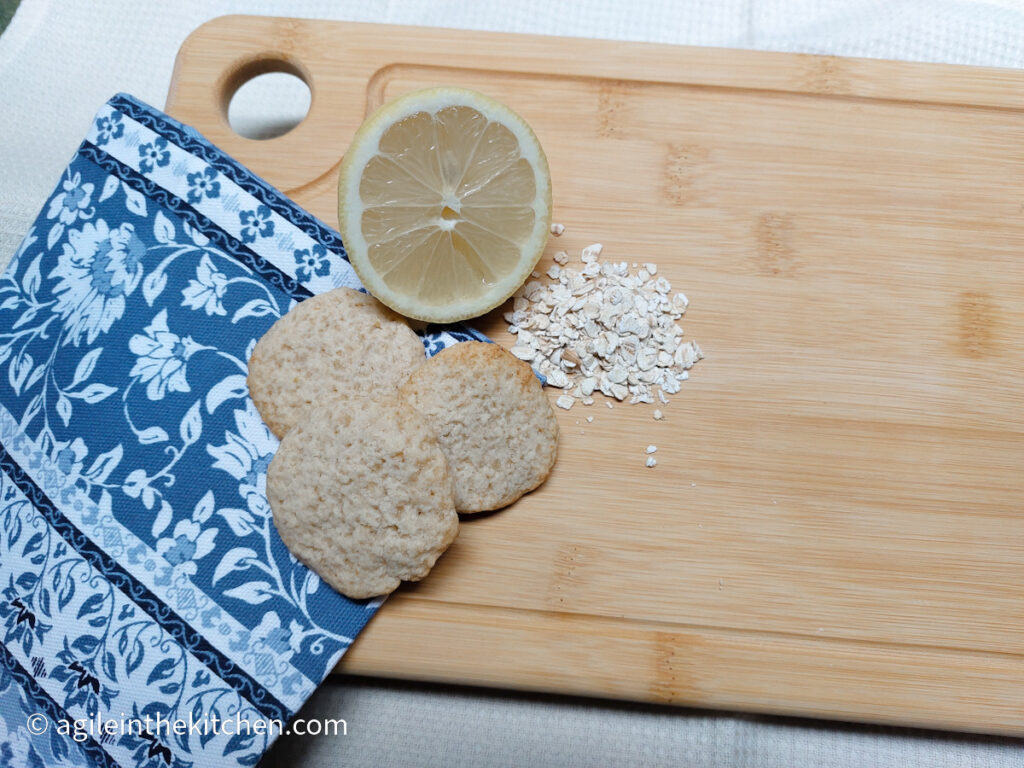 A wooden cutting board, with a folded blue coloured napkin with white flowers in the lower right corner. Three lemon oat cookies are stacked beneath a cut lemon, a handful of oats.