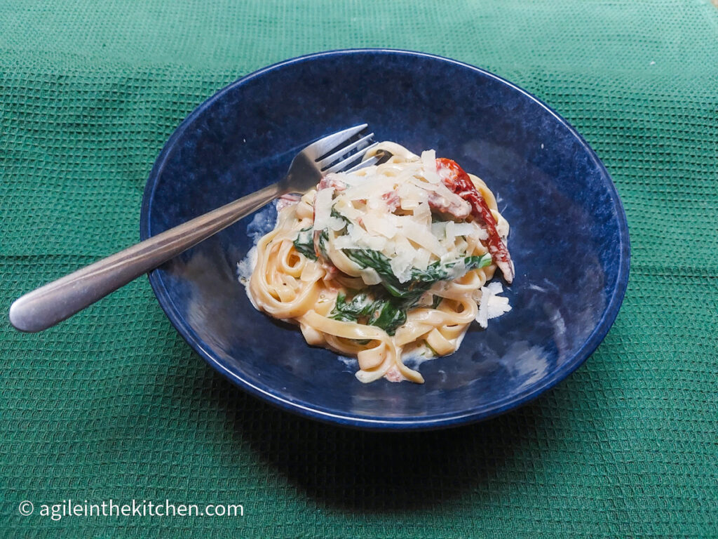 On a green table cloth, a blue bowl with tagliatelle and creamy sun dried tomato and spinach pasta sauce, topped with freshly grated Parmesan and a fork.