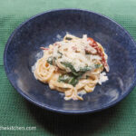 On a green table cloth, a blue bowl with tagliatelle and creamy sun dried tomato and spinach pasta sauce, topped with freshly grated Parmesan.