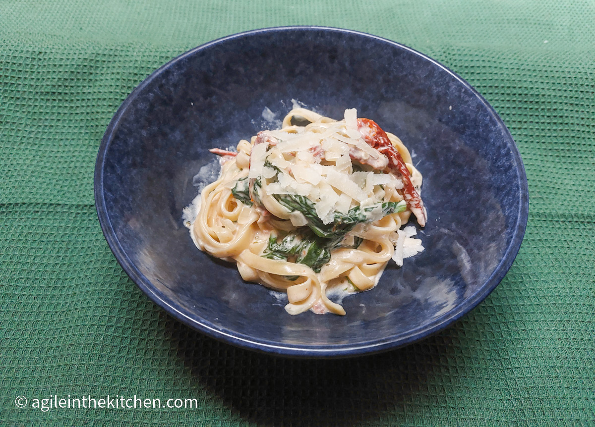 On a green table cloth, a blue bowl with tagliatelle and creamy sun dried tomato and spinach pasta sauce, topped with freshly grated Parmesan.