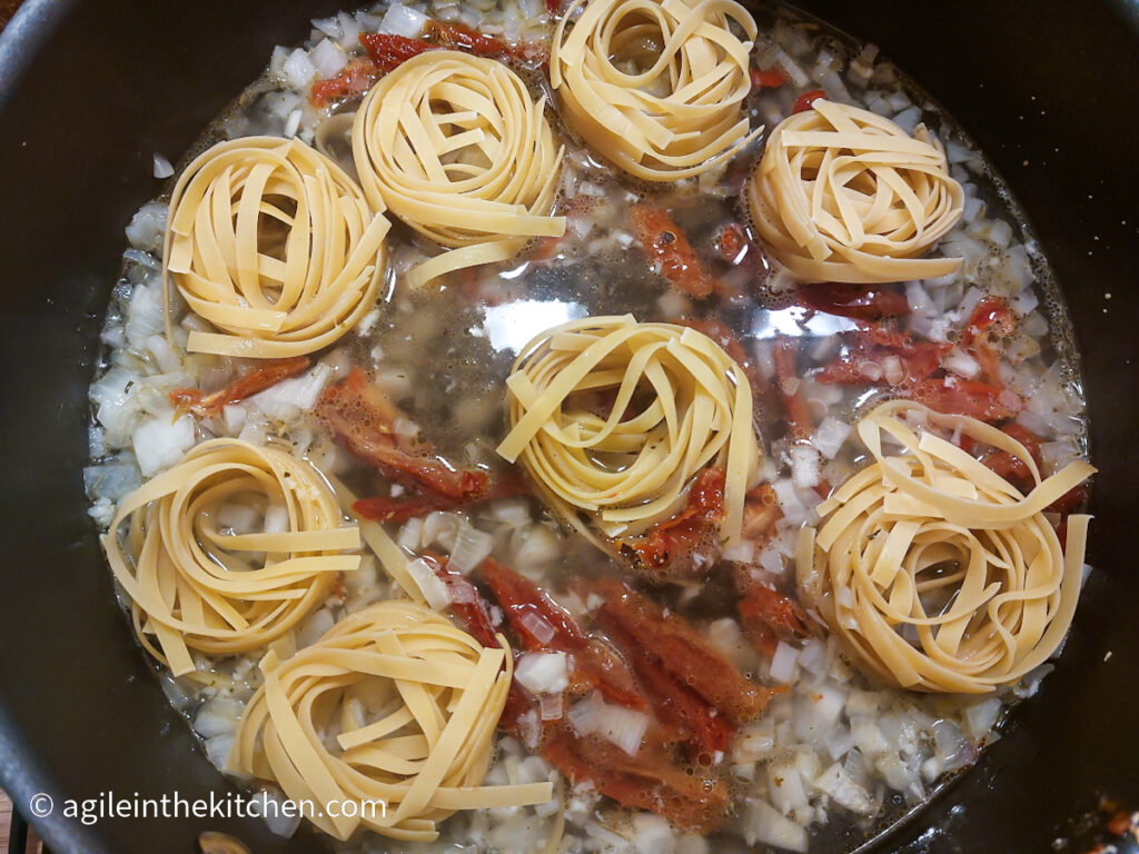 In a black pot tagliatelle nests are cooking with onion, sun dried tomatoes and water.