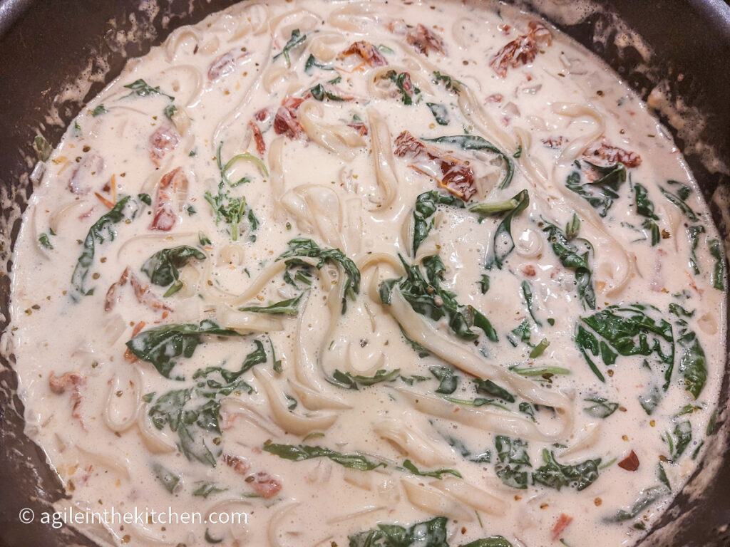 A large black pot the creamy sun dried tomato and spinach pasta is finishing cooking, visible in the creamy sauce is sun dried tomatoes, spinach and tagliatelle.