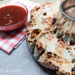 A close up of pull apart pizza bread and bbq sauce. On a beige textured background a red checkered napkin is folded, in the top left corner of the photo. On top of the napkin is a plate of bbq sauce. To the right of the napkin is a pull apart pizza bread, still half in its round oven form, all stuck together.