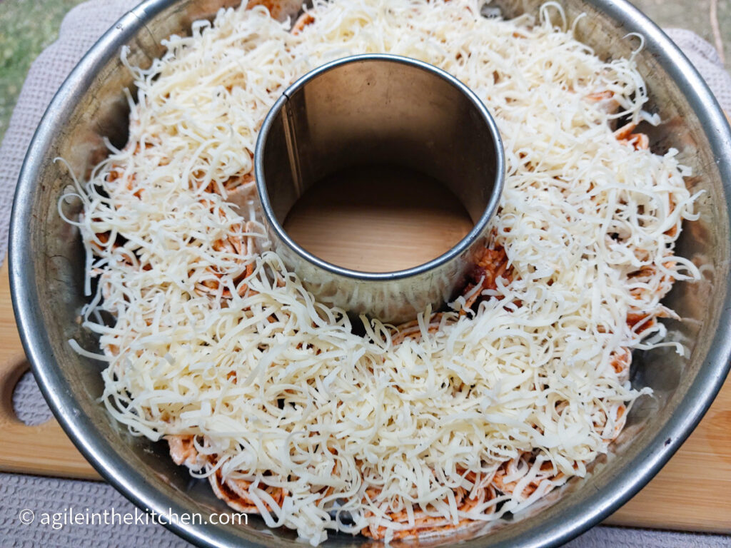 The photo taken from above, a pull apart pizza is complete with shredded cheese on top, sitting in its circular dish with a hole in the middle, on a wooden cutting board.